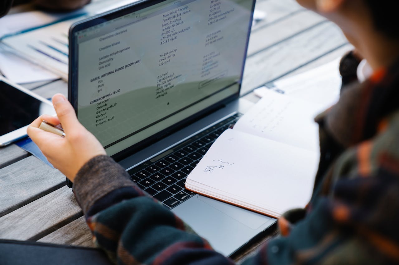 Close-up of hands using a laptop and taking notes at a wooden table.