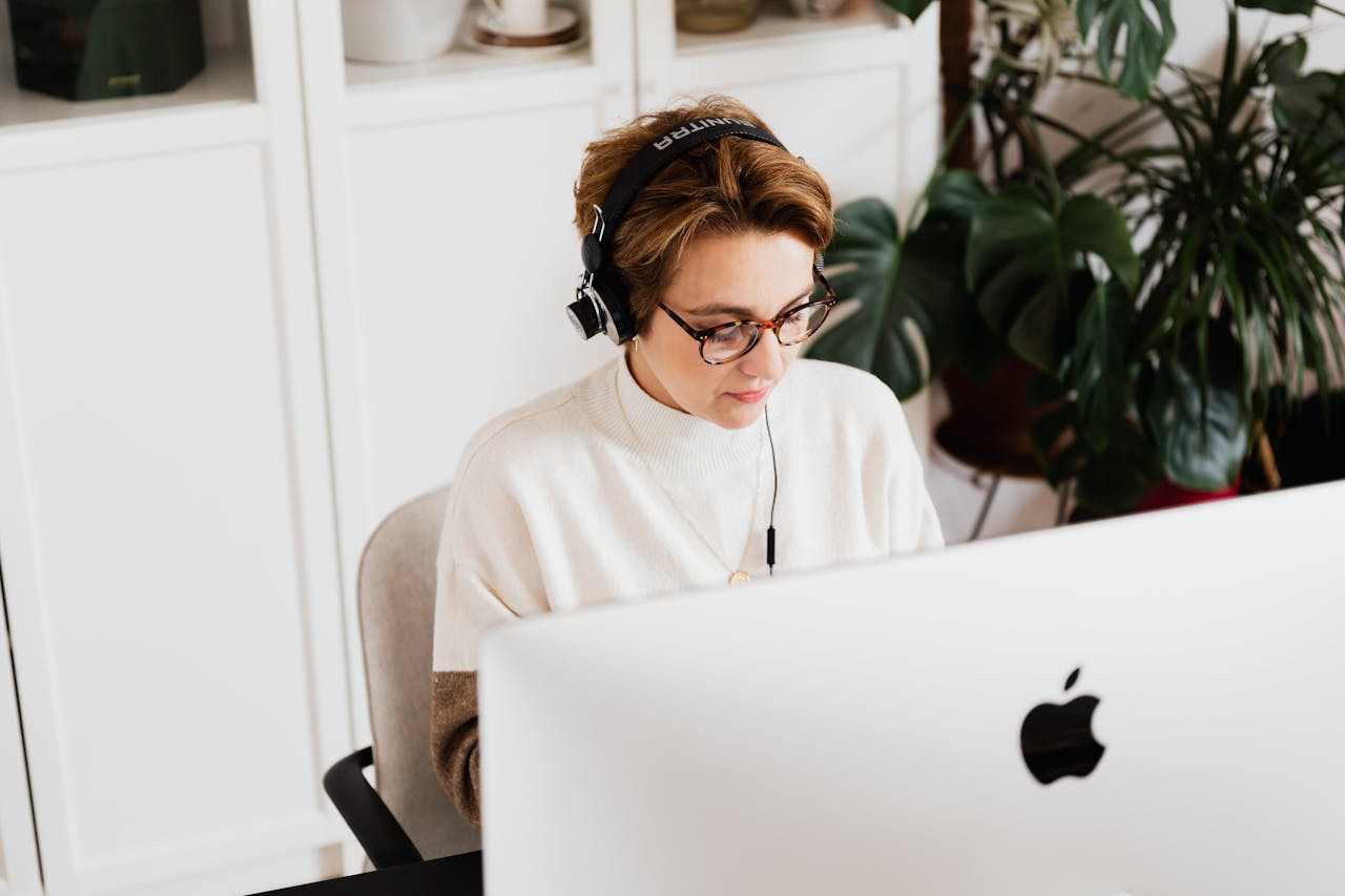 From above serious female in casual wear with eyeglasses and wired headphones using modern computer in contemporary light workplace